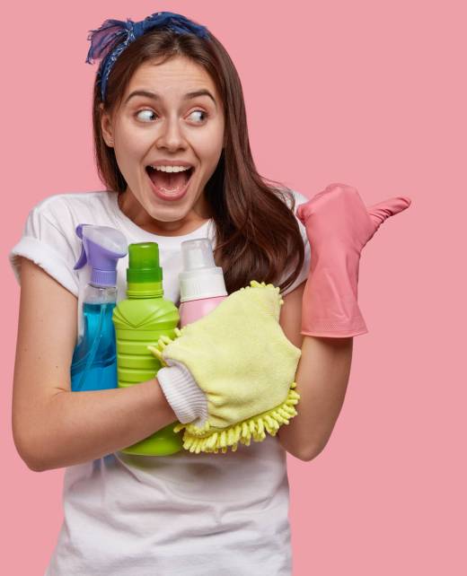 Glad Caucasian woman with positive expression, wears rubber gloves, points aside with thumb, holds spray and detergent in hands, dressed in casual white t shirt, models over pink studio wall